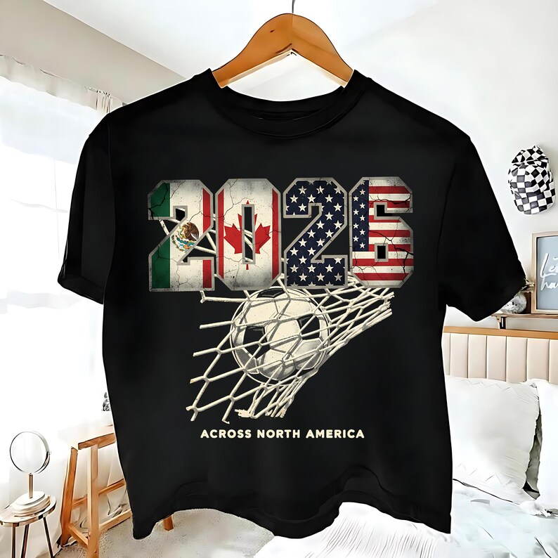 Op de afbeelding: Zwart t-shirt met het jaartal "2026", waarin de vlaggen van Mexico, Canada en de Verenigde Staten zijn verwerkt. Een voetbal zit in een net, met de tekst "ACROSS NORTH AMERICA" eronder.