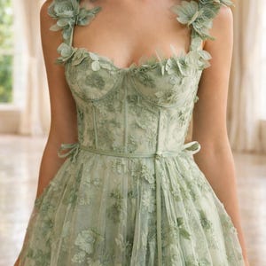 Puede incluir: Un vestido verde claro bordado con flores, con escote coraz&oacute;n y corpi&ntilde;o ajustado. El vestido presenta delicadas aplicaciones florales en los hombros y una falda fluida. Un cintur&oacute;n a juego ci&ntilde;e la cintura, y el vestido est&aacute; hecho de una tela transparente y en capas.