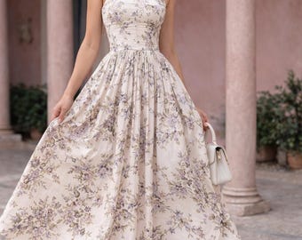 Puede incluir: Un vestido crema con estampado floral con un corpiño ajustado y una falda fluida. El vestido presenta un delicado estampado de flores moradas y verdes. El vestido tiene tirantes finos y se combina con un bolso blanco y sandalias beige. El fondo incluye columnas rosas.