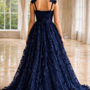 Puede incluir: Un vestido de noche azul marino con adornos florales. El vestido presenta un corpi&ntilde;o ajustado, tirantes finos y una falda amplia y fluida. Acentos florales adornan los hombros y caen por la falda. El vestido largo es adecuado para ocasiones especiales.
