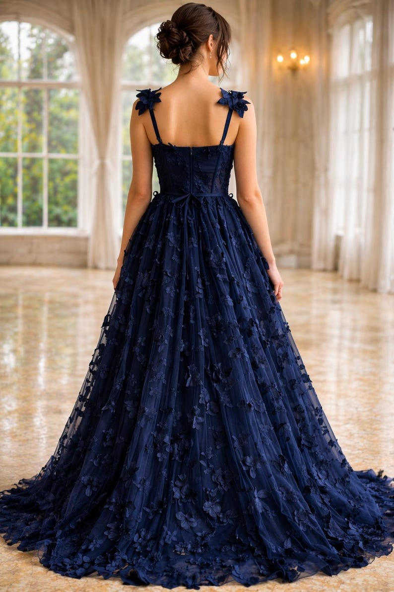 Puede incluir: Un vestido de noche azul marino con adornos florales. El vestido presenta un corpi&ntilde;o ajustado, tirantes finos y una falda amplia y fluida. Acentos florales adornan los hombros y caen por la falda. El vestido largo es adecuado para ocasiones especiales.