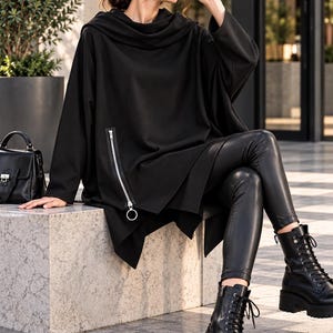 May include: A black hooded tunic featuring a long diagonal silver zipper with a circular pull, paired with black faux leather leggings and black lace-up combat boots. A small black handbag is positioned nearby.