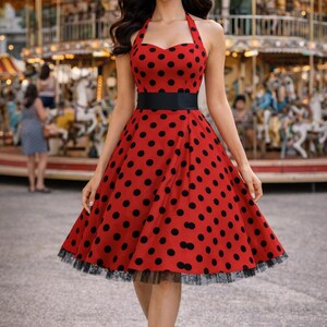 K&ouml;nnte beinhalten: Rotes Kleid mit schwarzen Polka Dots und schwarzem G&uuml;rtel. Das Kleid hat einen Neckholder-Ausschnitt und einen ausgestellten Rock mit schwarzem Spitzensaum. Im Hintergrund ist ein Karussell zu sehen.