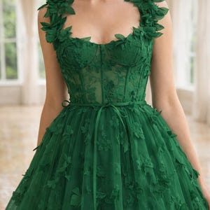 Puede incluir: Vestido de fiesta verde esmeralda con detalles florales. El corpi&ntilde;o presenta un escote coraz&oacute;n y un dise&ntilde;o de cors&eacute; ajustado. La falda es de tul amplio y fluido, adornada con aplicaciones florales en 3D. Una faja a juego ci&ntilde;e la cintura.