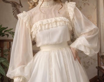 Puede incluir: Un vestido vintage de color marfil con un cuello alto con ribete de encaje y mangas largas transparentes. El vestido presenta delicados detalles de encaje en el corpiño y los puños, con una falda fluida y un cinturón de cinta en la cintura. El vestido recuerda a la moda victoriana.