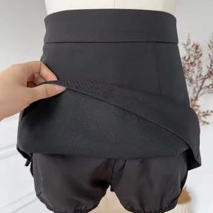 May include: Black skirt with attached shorts. The skirt has a high waist and a layered design, with the shorts visible underneath. The fabric appears to be a smooth, solid black material. The garment is displayed on a mannequin.