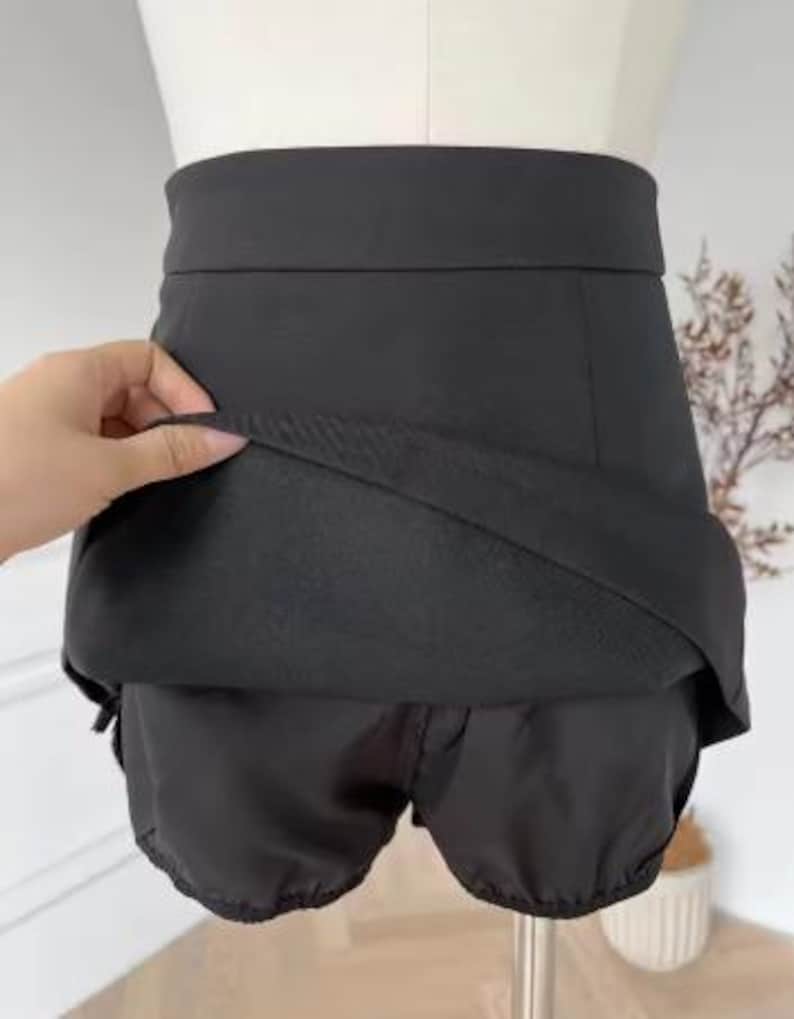 May include: Black skirt with attached shorts. The skirt has a high waist and a layered design, with the shorts visible underneath. The fabric appears to be a smooth, solid black material. The garment is displayed on a mannequin.