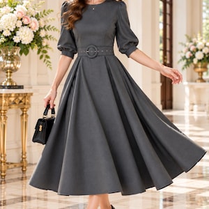 May include: A charcoal gray midi dress featuring a round neckline, elbow-length puff sleeves, and a matching fabric belt with a circular buckle at the waist. The skirt is pleated and flares out from the waist, and a small black handbag is held.