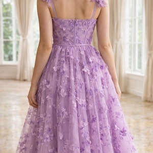 Puede incluir: Un vestido formal de color lavanda con adornos florales. El vestido presenta un corpi&ntilde;o ajustado, tirantes finos y una falda amplia hasta la rodilla. Los acentos florales adornan los hombros y se dispersan sobre la superposici&oacute;n de tul, creando un look rom&aacute;ntico y elegante.