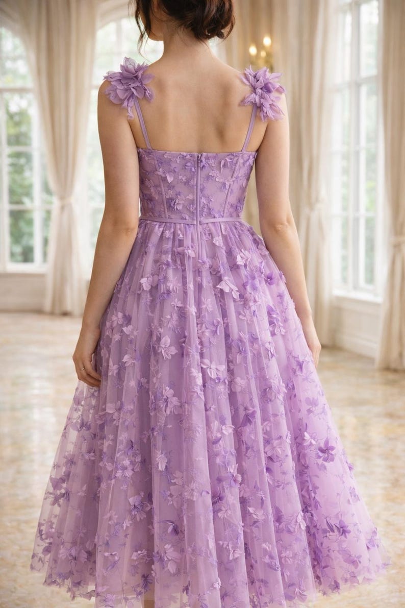 Puede incluir: Un vestido formal de color lavanda con adornos florales. El vestido presenta un corpi&ntilde;o ajustado, tirantes finos y una falda amplia hasta la rodilla. Los acentos florales adornan los hombros y se dispersan sobre la superposici&oacute;n de tul, creando un look rom&aacute;ntico y elegante.