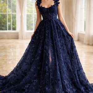 Puede incluir: Un vestido de noche azul marino con adornos florales. El vestido presenta un corpi&ntilde;o ajustado, una falda fluida y delicados detalles florales en los hombros y en la falda. El vestido tiene una abertura alta y una larga cola.