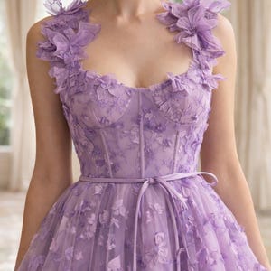 Puede incluir: Un vestido formal de color lavanda con adornos florales. El corpi&ntilde;o presenta un dise&ntilde;o tipo cors&eacute; y est&aacute; adornado con delicadas flores de tela. El vestido tiene una falda fluida y una cinta en la cintura a juego. Acentos florales tambi&eacute;n en los hombros.
