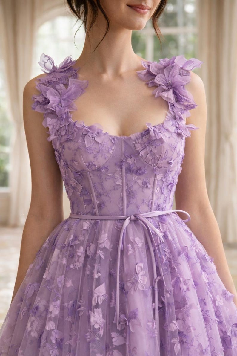 Puede incluir: Un vestido formal de color lavanda con adornos florales. El corpi&ntilde;o presenta un dise&ntilde;o tipo cors&eacute; y est&aacute; adornado con delicadas flores de tela. El vestido tiene una falda fluida y una cinta en la cintura a juego. Acentos florales tambi&eacute;n en los hombros.