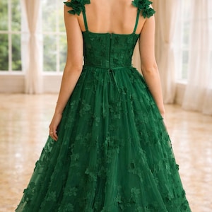 Puede incluir: Vestido de noche verde esmeralda con adornos florales. El vestido presenta un corpi&ntilde;o ajustado, tirantes delicados y una falda de tul fluida adornada con aplicaciones florales en 3D. La espalda tiene cierre de cremallera y lazo.