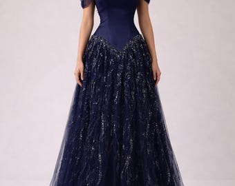 Puede incluir: Un vestido de noche azul marino con un corpiño ajustado y una falda amplia y fluida. El corpiño presenta mangas descubiertas y un cuello choker. La falda está adornada con brillantes detalles plateados, creando un look glamuroso y elegante.