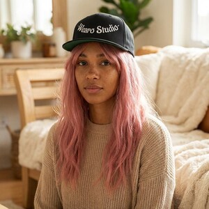 Op de afbeelding: Zwarte baseballcap met de tekst "Wearo Studio" in witte letters. De cap heeft een groene klep. De persoon op de foto draagt een beige trui en heeft roze haar.
