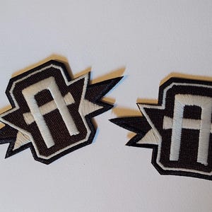 Puede incluir: Dos parches bordados con la letra "A" blanca sobre fondo marrón, enmarcados por un diseño de estrella negra y blanca. Los parches son adecuados para ropa o accesorios.