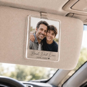Op de afbeelding: Transparante acryl auto zonneklep fotohouder met een foto van een vader en zoon. De tekst "Best Dad Ever" staat onder de foto, samen met "LOVE, JAMES" en een hartsymbool. De zonneklep is beige.