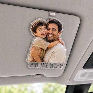Peut inclure: Ornement de pare-soleil de voiture en acrylique transparent avec une photo d'un père et d'un enfant souriants s'embrassant. L'ornement a un bord incurvé et le texte "DRIVE SAFE DADDY" en lettres noires, avec un symbole de cœur.