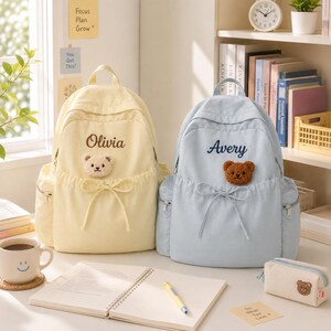 Pode incluir: Uma mochila amarelo-pálido com o nome Olivia e um pequeno emblema de urso creme, e uma mochila azul-clara com o nome Avery e um emblema de urso castanho. Ambas as mochilas apresentam um detalhe de cordão frontal, bolsos laterais e fechos prateados.