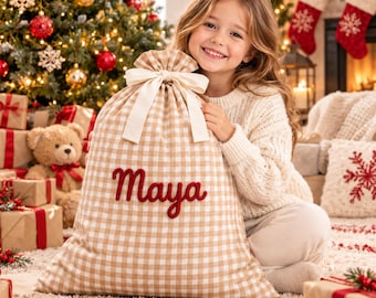May include: A large, beige and white gingham fabric gift sack featuring a drawstring closure with a cream-coloured ribbon bow. The name Maya is embroidered in red script lettering on the front of the sack, which is made of a woven textile.