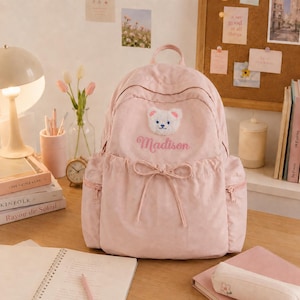 Könnte beinhalten: Ein hellrosa Rucksack mit einem flauschigen weißen Bärengesicht-Aufnäher, dem gestickten Namen Madison und einer dekorativen Kordelschleife an der Vordertasche. Der Rucksack verfügt über zwei Seitentaschen mit Reißverschlüssen und verstellbare Träger.