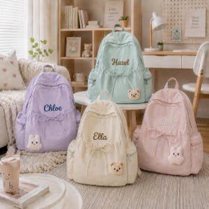 Op de afbeelding: Vier rugzakken in pastelkleuren: lavendel, mintgroen, crème en lichtroze. Elke tas heeft een trekkoordsluiting, zijzakken, een pluche dierenpatch en geborduurde namen: Chloe, Hazel, Ella en Ruby in een cursief lettertype.