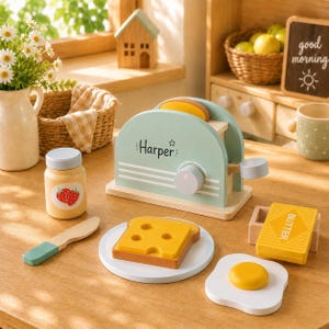 May include: A wooden toy toaster in sage green with the name Harper printed on the side, accompanied by a wooden slice of toast, a yellow butter block, a fried egg, a strawberry jam jar, and a butter knife, all arranged on a wooden surface.