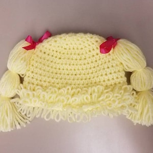 May include: A yellow crocheted hat with two long yarn pigtails and pink bows. The hat has a fringe around the bottom.