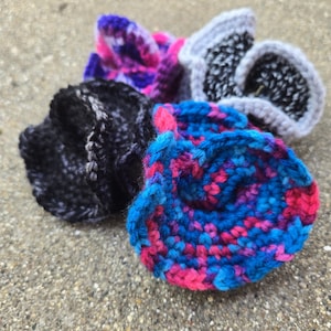 May include: Four handmade crochet scrunchies in shades of blue, pink, purple, gray, and black. The scrunchies are made of soft yarn and have a textured appearance. The scrunchies are in a variety of patterns and colors.