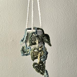 May include: A handmade crochet plant hanger with a white pot and hanging leaves. The leaves are in shades of green and blue. The hanger is suspended by three white cords.