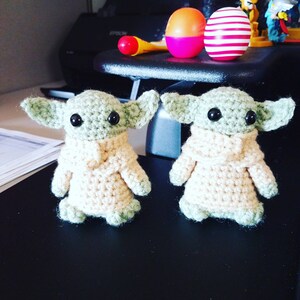 May include: Two crocheted green and beige baby Yoda figures. The figures are standing on a black surface.