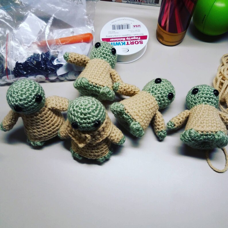May include: Five crocheted green and beige baby Yoda figures with black button eyes. The figures are sitting on a white surface.