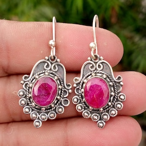 Puede incluir: Un par de pendientes colgantes con piedras preciosas de color rojo rosado intenso de corte ovalado engastadas en plata oxidada. El metal presenta intrincados patrones de volutas, bordes de cuerda trenzada y acentos esféricos.