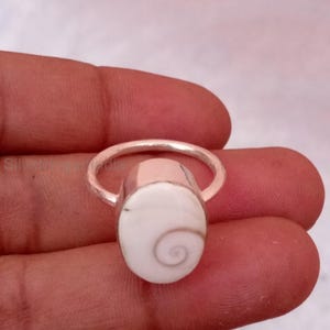 Könnte beinhalten: Ein silberner Ring mit einem ovalen, weißen Stein mit einem Spiralmuster. Der Ring hat ein schlichtes Band und eine Fassung, die die Form des Steins hervorhebt. Der Ring wird in einer Hand gehalten, um sein Design und seine Details zu zeigen.