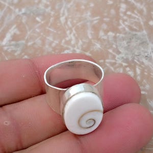 May include: A silver ring featuring a smooth, polished band and a prominent oval-shaped white shiva eye shell inlay. The shell displays a natural, spiraling brown pattern centered on its surface, securely set within a simple silver bezel setting.