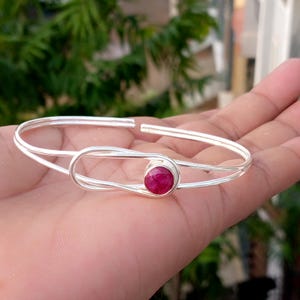 Puede incluir: Una pulsera de brazalete de plata con un diseño de banda abierta pulida y una piedra preciosa circular facetada de color rosa intenso, engastada en un bisel de plata liso. El metal tiene un acabado brillante y una estructura sencilla.
