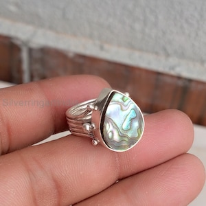 May include: A silver ring featuring a teardrop-shaped abalone shell inlay with iridescent green, blue, and purple tones. The band is constructed from multiple thin, stacked silver wires, accented with small silver spheres at the setting base.