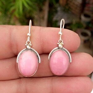 May include: A pair of silver drop earrings featuring smooth, oval-shaped pink stones set in polished metal frames. Each earring hangs from a simple silver hook, showcasing a minimalist design with a rounded, cabochon-cut pink gemstone centerpiece.