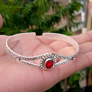Puede incluir: Una pulsera de plata con diseño abierto y patrones grabados intrincados a lo largo de la banda. El centro de la pulsera muestra una única piedra preciosa de color rojo intenso y forma ovalada engastada en un marco de plata decorativo.