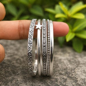 Peut inclure: Un ensemble de cinq bracelets joncs argentés aux textures et motifs variés. Un bracelet présente un motif de vigne florale, un autre arbore une petite breloque étoile, tandis que les autres affichent des surfaces géométriques, perlées et lisses.
