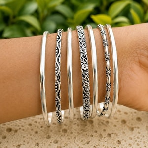 Op de afbeelding: Een set van zes zilverkleurige armbanden die samen worden gedragen. De collectie bevat twee gladde, gepolijste banden, één met een golvend lijnpatroon, één met een herhalend bloemmotief, één met een kralentextuur en één met een ruitvormig ontwerp.