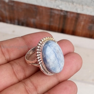 May include: A silver ring featuring a large, oval-shaped, light blue cabochon stone. The setting is decorated with a beaded metal border around the stone. The band is made of polished silver and includes a visible 925 hallmark stamp on the side.