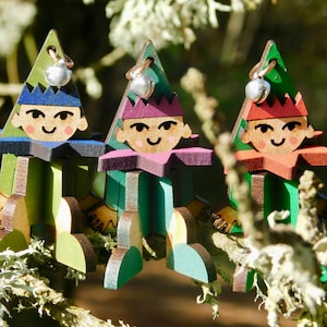 May include: Three wooden elf decorations with smiling faces, each with a different coloured hat and collar. The elves are green, blue, and red, and each has a silver bell at the top. The decorations are displayed on a tree branch.