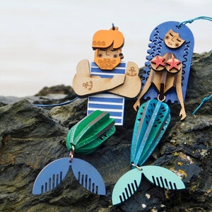 May include: Two wooden ornaments depicting a mermaid and a sailor. The mermaid is blue with red stars on her chest and a teal tail. The sailor is wearing a blue and white striped shirt and has a beard and a tattoo. Both ornaments are hanging from a string.