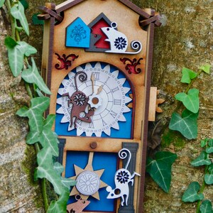 May include: A decorative wooden cuckoo clock with a mouse theme. The clock has a light brown frame with blue, red, and white details. The clock is hanging on a tree trunk with green ivy. The clock's measurements are in centimeters.