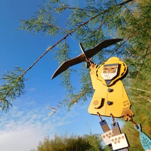 May include: A yellow wooden figurine of a person wearing a raincoat and holding a bird. The figurine is hanging from a tree branch against a blue sky.