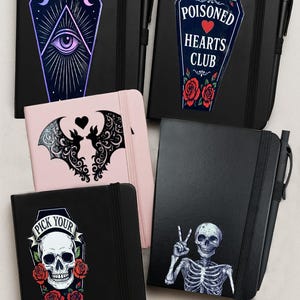Puede incluir: Una colección de cuadernos negros con diseños únicos. Uno presenta un ataúd con un ojo y lunas crecientes, otro dice "Poisoned Hearts Club", y un tercero tiene una calavera con rosas. Cada cuaderno incluye un bolígrafo.
