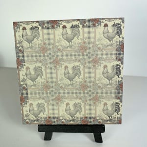 May include: A decorative square print featuring a repeating pattern of roosters, roses, and plaid. The print is in shades of cream, red, gray, and black. The print is displayed on a small black easel.