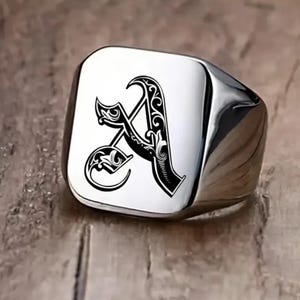 Könnte beinhalten: Ein silberner Siegelring mit quadratischer Oberfläche. Der Ring zeigt einen schwarzen, verzierten Buchstaben "A" in der Mitte. Der Ring hat eine polierte, reflektierende Oberfläche.